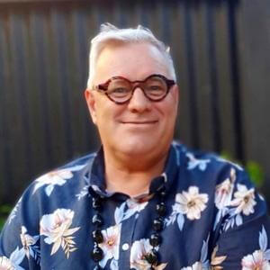 Warren Elwin headshot wearing flowery shirt and ula necklace
